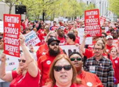 Rows and rows of people marching outside all with red shirts on and holding signs about Schools