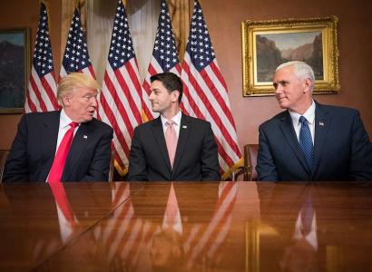 Trump, Ryan and Pence at a table looking at each other with flags in the back