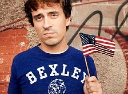 Young brown haired man wearing a blue t-shirt saying Bexley and waving a tiny flag in front of some kind of wall with words on it
