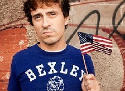 Young  white man  with tousled brown hair wearing a blue T-shirt that says BEXLEY in white letters and he's waving a very tiny flag and kind of sneering at the camera in a bored way