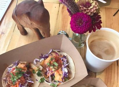 A top down view of a meal - a brown holder with what looks like two taco-like things, and brown drink in a white cup, something in a brown holder at the bottom, white potatoes with something green sprinkled on top and a red flower in a vase and an elephant statue