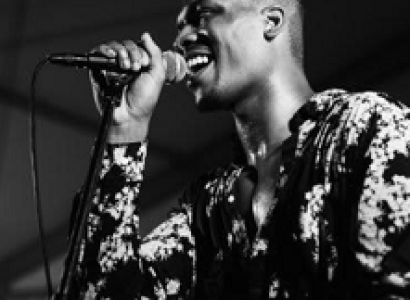 Black and white photo of bald black man smiling and singing into a mic with a flowered shirt on