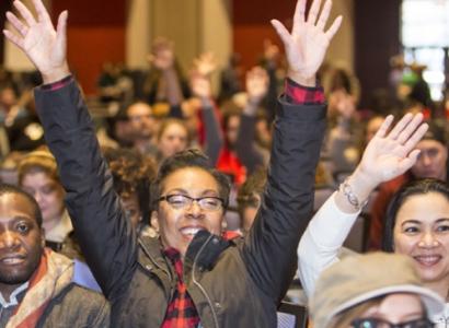 Black woman with glasses in front of a huge crowd of people smiling with both hands held high above her head