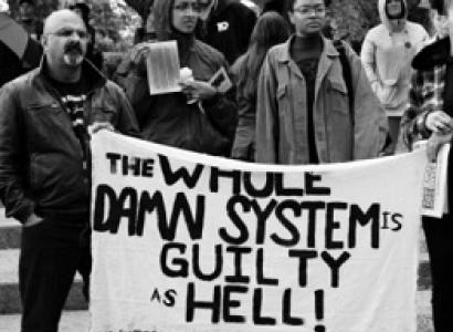 Black and white photo with people in winter coats outside gathered in a group behind a white banner with black letters saying The Whole Damn System is Guilty as Hell