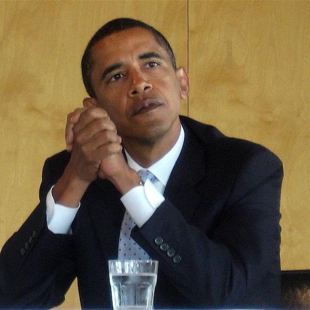 Obama with elbows on table clasping his hands together looking pensive
