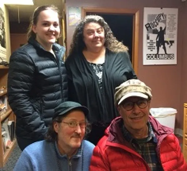 Two women, one young with hair pulled back and the other older with long brown curly hair standing behind two men kneeling, both wearing hats with brims and wire rimmed glasses, everyone smiling