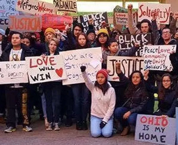 Many people posing in rows outside wearing winter clothes holding signs that say Live Will Win, Refugees Welcome, Justice, Ohio is my Home, Solidarity, Organize, and a woman in front on her knees with a fist in the air