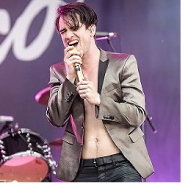 Young white man with satiny suit jacket and no shirt with shaggy brown hair singing intoa ic