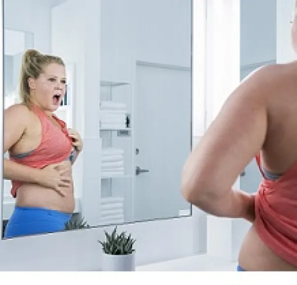 A blond woman with her hair in a knot of top of her head looking out as an image in a mirror with her mouth open and showing her belly between a red bra top and blue bottom with her hand on her belly