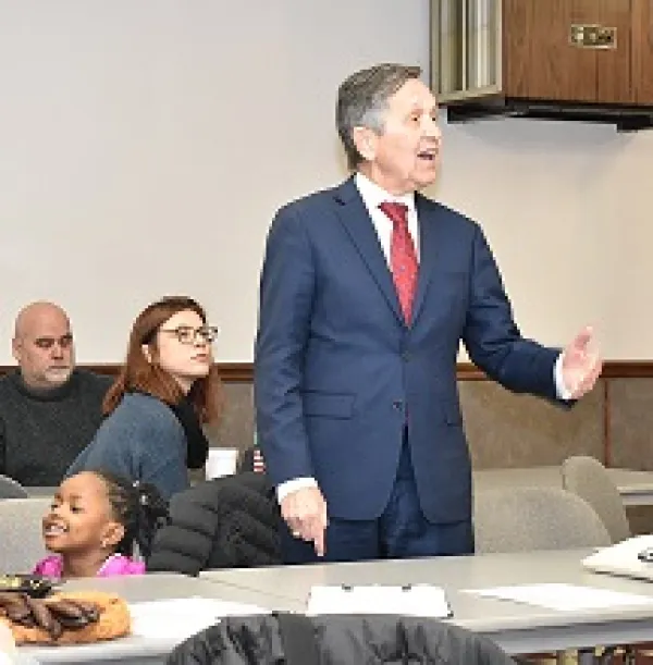 Middle aged white man with some dark and some gray hair speaking while gesturing to the right wearing a suit, people listening to him