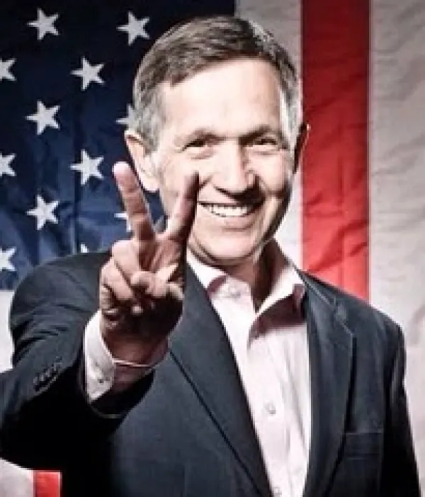 Older man with gray sideburns smiling and holding his fingers in a peace sign with a flag in the background