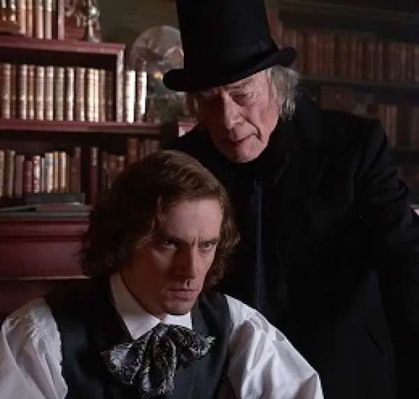 A man with chin length curly brown hair looking intense sitting down with old fashioned clothes from the 30s and an older gray haired man in black with a black top hat leaning over his shoulder saying something in his ear, bookshelves in the background