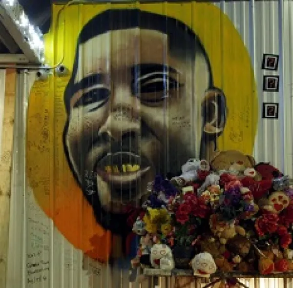 Painting of a black man's face against a yellow and orange circle on a white sided building with a bouquet of flowers in the foreground
