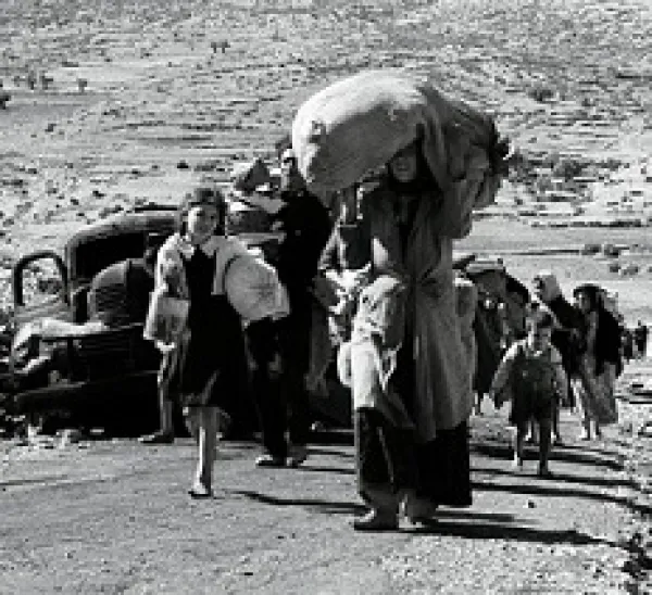 Black and white photo of women and children walking down a dusty path outside with the woman in the foreground carrying a big heavy bag on her head