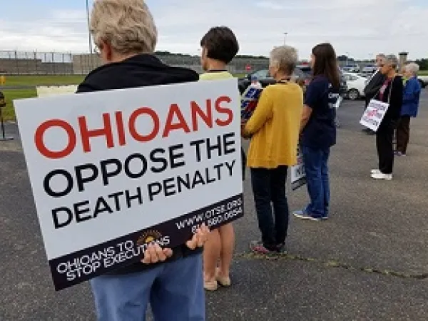 The backs of four people at a vigil outside one holding a sign that says Ohioans Oppose the Death Penalty