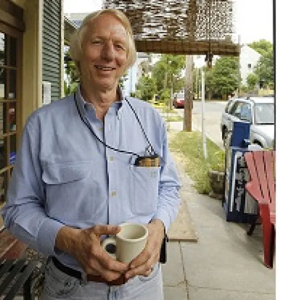 Thin tall white man with white hair smiling holding a coffee cup standing outside a store with a outdoor chair
