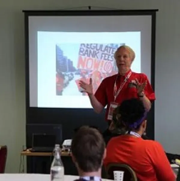 Gray haired man in red shirt waving his arms in front and talking to a group seated before him with a projected image on a screen behind him