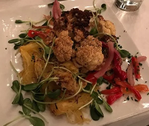 A white square plate on a table filled with greens, little toasty looking brownish balls, squash and red slivers of food