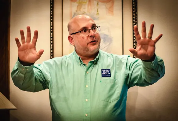 White middle aged guy with glasses holding his arms in the air with fingers spread on hands while he talks and wears an Abolish the Death Penalty button