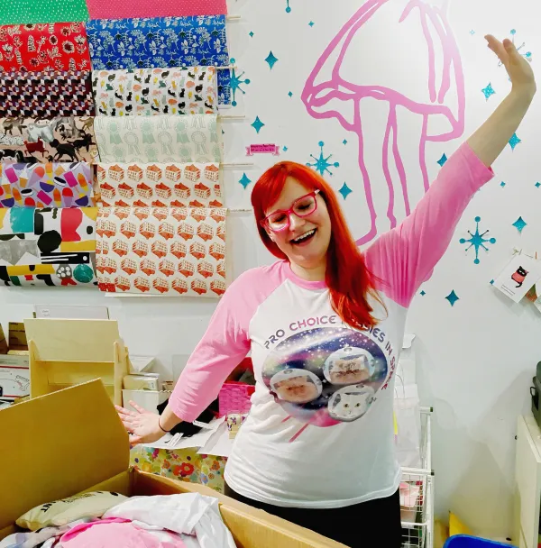Woman with red hair and pink glasses holding up her hand like waving hello in a room with a lot of arts and crafts supplies