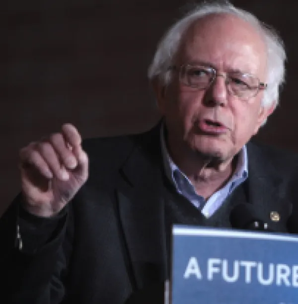 Older white man with white hair and wire rimmed glasses gesturing and speaking at a podium