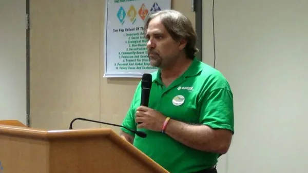 White man with goatee and dark hair in green shirt holding a mic at the podium