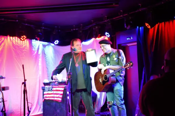 Two men on a stage, one with a guitar, one with a plaque in his hand