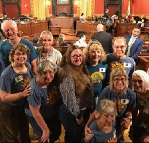 Lots of smiling white people male and female leaning toward each other all wearing blue T-shirts in a city council chambers