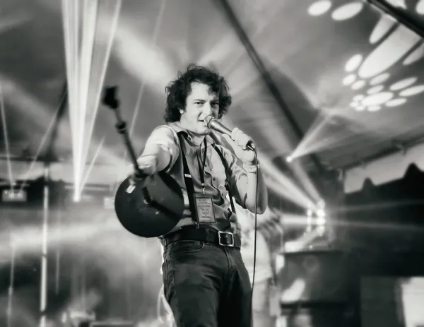 Black and white photo of young dark haired man singing into a mic on a stage
