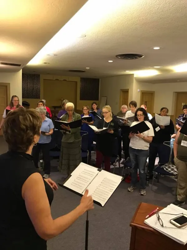 Lots of women in a half circle singing with one woman conducting
