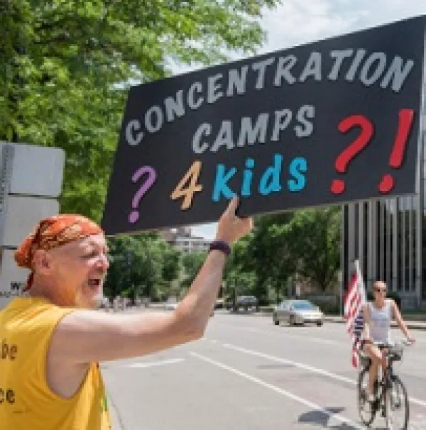 White man in a bandanna and yellow shirt yelling and holding a sign that reads Concentration Camps 4 Kids with lots of question marks