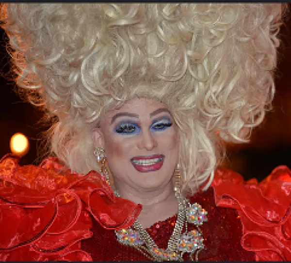Man dressed a woman with huge blonde hair in curls going up in the air lots of blue eye shadow, red lips and a red outfit