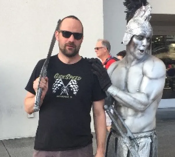Guy with dark beard, sunglasses and sword next to a silver statue of a half-naked warrior with metal helmet