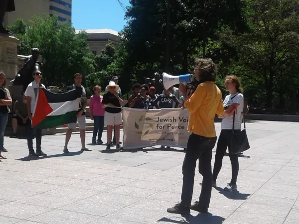 Man speaking into bullhorn and crowd with Jewish Voices for Peace sign