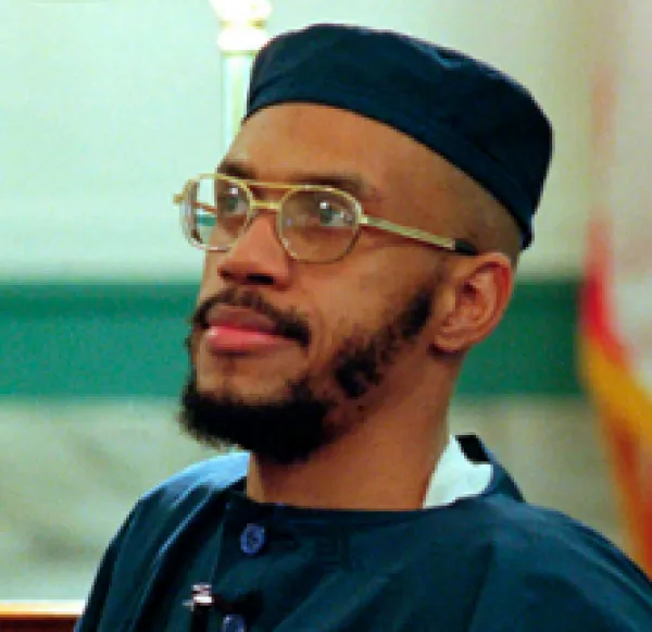 Black man with wire rimmed glasses and mustache and beard and a square topped blue hat