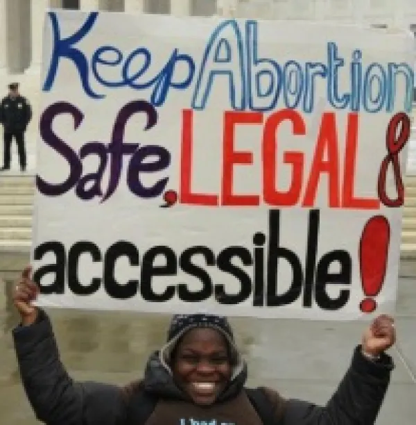 Black woman holding sign saying Keep abortion safe, legal and accessible in from of a government building with a cop standing in the background