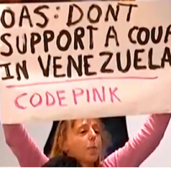 Blonde woman in pink shirt holding big sign above her head that says OAS: Don'ts support a coup in Venezuela