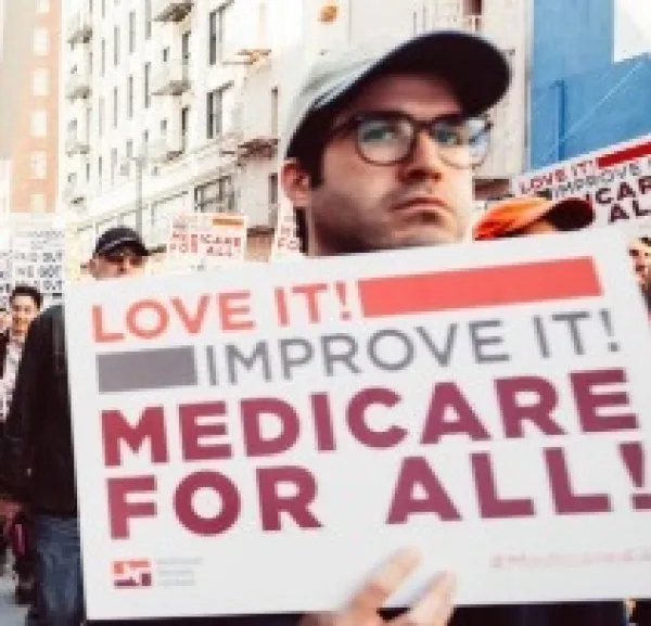 White man in a baseball cap and glasses holding a sign that says Love It Improve It Medicare for All