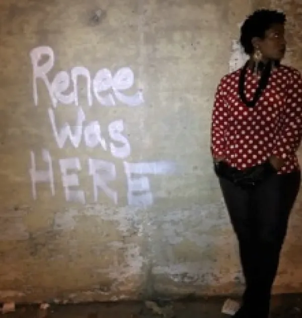 The words Renee was HERE spray painted on a brown wall and a black woman with short hair and a red and white shirt with a black necklace and black pants stands against the wall looking to the right