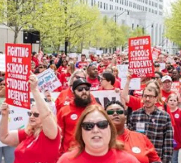 Rows and rows of people marching outside all with red shirts on and holding signs about Schools