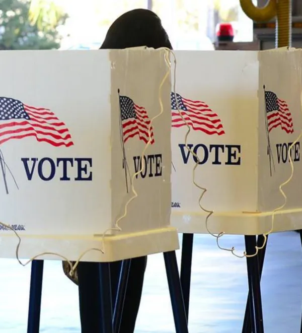 Person in voting booth that is white with a flag on the back and word VOTE