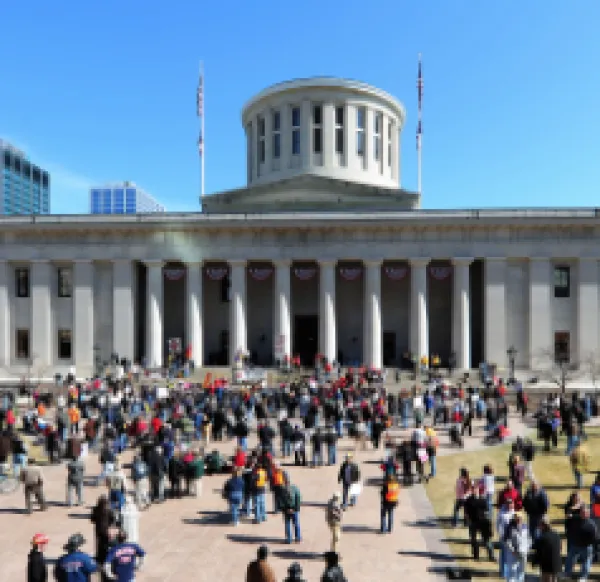 Big government building with round tower at top and white columns and lots of people on the grounds in front
