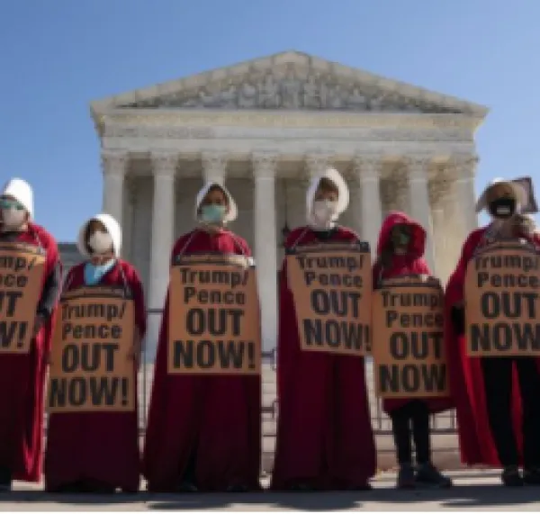 Proestors dressed like handmaidens outside a government building with signs saying Trump Pence Out Now
