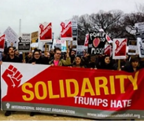 Lots of people at a march holding picket signs and a very large long banner with red fist and words Solidarity trumps hate
