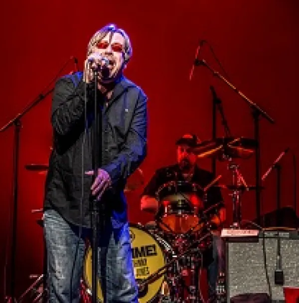 Middle aged white man with red sunglasses on singing with wide mouth open into a standing mic in front of a guy playing drums