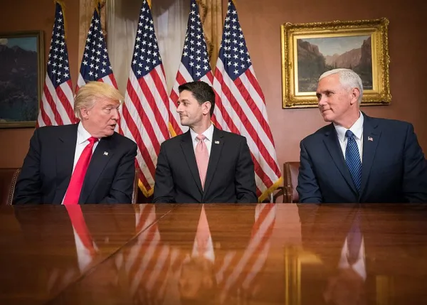 Trump, Ryan and Pence at a table looking at each other with flags in the back