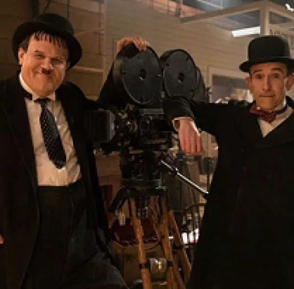 One large and one small man both in black suits with white shirts and hats leaning on an old time film projector