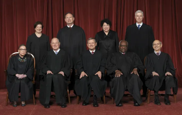 Photo of men and women posing and facing the camera all wearing long black robes in front of a dark red curtain