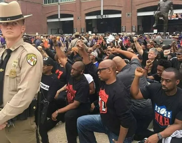 Lots of black men down on one knee with fists raised in the street with police man in foreground