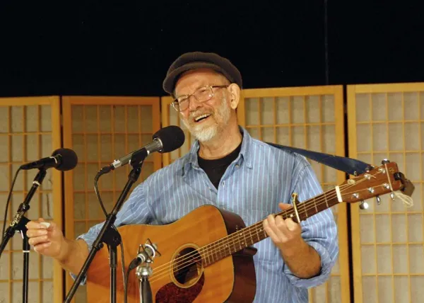 Man with gray goatee and black cap holding guitar at microphone smiling 